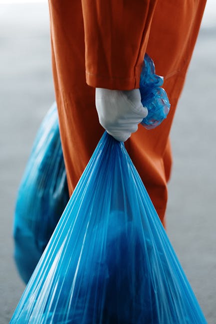 A close-up view of a person dressed in orange overalls, wearing a white glove, holding a large, tightly tied blue plastic rubbish bag by the top. The bag appears to be filled with waste materials and is slightly translucent, revealing some contents inside. The background is blurred but suggests an outdoor environment, possibly near a waste disposal site or a street. The individual is positioned on the right side of the image, with the bag extending downward and slightly to the left. This scene exemplifies a thorough rubbish collection process, corresponding to independent waste handling or on-site clearance services provided by Rubbish Removal Croydon, highlighting the practical aspects of rubbish collection and disposal in a professional and neutral manner.