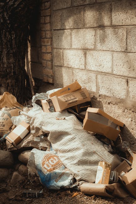 An outdoor scene showing a pile of miscellaneous rubbish against a brick wall, including cardboard boxes in various sizes, some partially flattened, and a white woven sack contaminated with dirt and debris. The boxes have visible printed labels and are open or crushed, with edges bent and torn, indicating recent disposal or collection. The sack is positioned on rough ground composed of small stones and soil, partially covering other waste materials. In the background, there is a weathered brick wall and a section of a tree trunk, suggesting the rubbish is situated in an external area adjacent to a building or garden. The lighting is natural, casting soft shadows over the debris, emphasizing the textures of the materials. This scene reflects private or independent rubbish disposal efforts, aligning with non-council waste collection services such as those offered by Rubbish Removal Croydon, focusing on effective on-site clearance of domestic or small commercial waste.