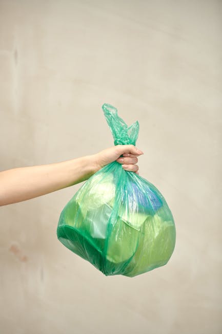A person’s hand and forearm are visible in the image, holding a semi-transparent green plastic rubbish bag tightly by its twisted top, which is gathered and knotted. The bag appears to be filled with waste, giving it a rounded, slightly bulging shape, with some hints of blue and other colours distorted by the plastic. The background is a plain, light beige or off-white wall, providing a neutral backdrop that highlights the bag and the hand. The lighting is even, with no strong shadows, emphasizing the clarity of the plastic material and the details of the hand’s skin. This scene depicts a typical handling of waste for disposal, relevant to private rubbish collection services, such as those offered by Rubbish Removal Croydon, emphasizing the importance of proper waste containment before removal or disposal, especially in the context of independent rubbish collection projects or on-site clearance activities within residential or commercial environments.