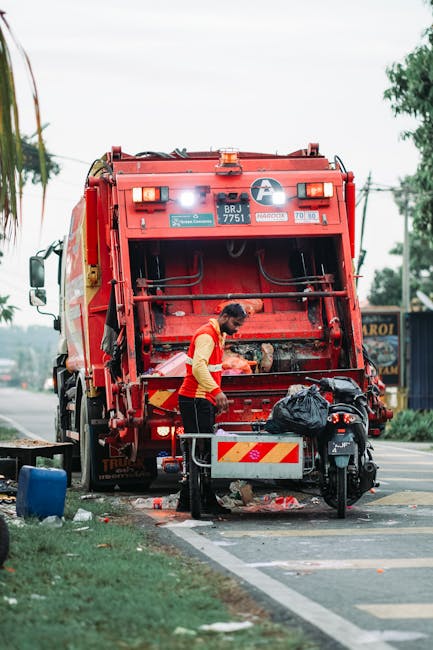A waste collection worker wearing a yellow and orange high-visibility vest is standing in front of a large red rubbish collection truck on a street. The truck, positioned with its rear opening facing the viewer, is equipped with mechanical components and safety markings, and it appears to be in the process of emptying or collecting waste. The worker is handling a black plastic bag, which is placed on a small trolley or bin attached to the truck. The scene includes a vehicle lane, a sidewalk, and some greenery on the side of the street, with trees and power lines visible in the background. The environment is outdoors, during daylight hours, and the overall setting demonstrates a typical urban or suburban waste collection operation, emphasizing private or independent rubbish removal services like those offered by Rubbish Removal Croydon.