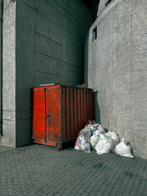 A weathered, red metal waste container with visible rust and chipped paint, positioned against a concrete wall at the corner of an urban alleyway. Several white and transparent plastic rubbish bags, filled and loosely tied, are placed on the ground next to the container, with some leaning against its side. The surrounding environment includes dark, smooth wall surfaces and a textured, paved ground made of interlocking bricks. The scene appears to be outdoors, possibly part of a private waste collection or rubbish removal service in Croydon, with natural diffused lighting contributing to a neutral, utilitarian atmosphere that emphasizes the practical aspect of waste disposal close to a commercial or residential area.