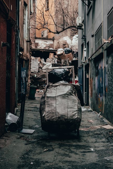 A narrow urban alleyway filled with a large collection of waste and rubbish. In the foreground, a large grey and black bag or sack sits on the dark, uneven pavement, which appears stained and littered with small debris. Behind the bag, there is a pushcart or trolley loaded with additional rubbish, including black plastic bags, cardboard boxes, and miscellaneous waste materials. The background features a cluttered heap of various waste, with uneven layers of cardboard, paper, plastic, and other refuse piled up against a wooden structure or fence. The alley is bordered on both sides by tall, weathered buildings with graffiti and peeling paint, and leafless tree branches extend overhead, suggesting a cold or winter season. Soft, diffuse lighting highlights the textures of the waste and the gritty urban environment, emphasizing the utilitarian nature of the scene. This image visually relates to private waste disposal services offered by companies like Rubbish Removal Croydon for efficient on-site clearance and alternative rubbish collection options near East Croydon station.