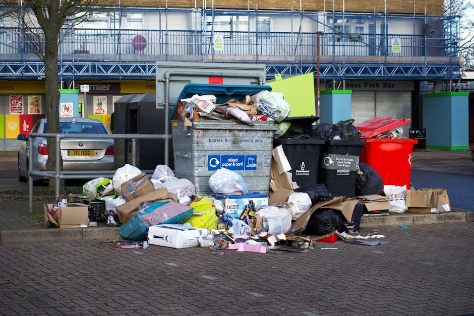 An outdoor scene showing an overflowing collection of mixed waste and rubbish bags situated on a paved parking area adjacent to a metal railing. The waste includes crumpled cardboard boxes, paper and plastic packaging, black garbage bags, and loose assorted debris; some items are spilling onto the street surface. Behind the pile, there are two green-labeled recycling bins, one black and one red, with partially visible logos, alongside a large grey waste container marked for mixed paper and card recycling. In the background, a parked car with a yellow registration plate is visible next to the pile, and a shopfront with yellow signage and closed shutters is seen further behind. The scene is outdoors under natural light, with a tree on the left side and a building with scaffolding and a blue safety barrier above, indicating possible ongoing maintenance or construction nearby. The image reflects a typical example of waste accumulation that might be handled through private rubbish removal services such as those offered by Rubbish Removal Croydon.