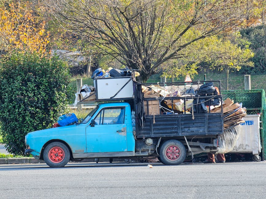 A small blue pickup truck parked alongside a street, loaded with a large amount of mixed rubbish and debris. The truck's rear bed is filled with cardboard boxes, plastic bags, and other waste materials, some secured with ropes. The vehicle has red-painted wheels and a slightly weathered exterior. Behind the truck, there is a green hedge and a tree with fresh green and yellow leaves, indicating an autumn setting. A large white container or bin is positioned at the rear right side of the truck, with a logo partially visible. The scene appears to be in a residential or semi-urban environment, with a fence and trees in the background. The overall impression is of an on-site or independent waste collection or rubbish removal effort, consistent with private disposal services such as those offered by Rubbish Removal Croydon.