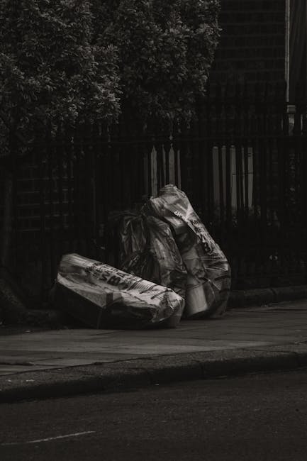Two large black plastic garbage bags filled with waste material are placed on the pavement in front of a black metal fence, which encloses a garden or yard area with thick foliage. The bags appear to be tightly knotted at the top and are positioned on the curb, suggesting a nearby rubbish collection service. The scene is outdoors, with a dark, overcast atmosphere, and the surrounding environment includes a strip of asphalt road and part of a residential or commercial building in the background. The image highlights the act of private rubbish disposal, typical in the context of alternative waste handling or on-site clearance facilitated by a private company such as Rubbish Removal Croydon, used to manage household or commercial waste outside of local authority collection.