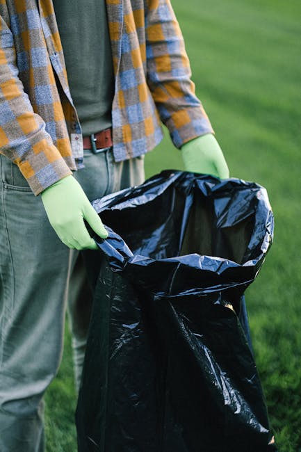 A person stands outdoors on a grassy area, wearing a patterned jacket with yellow, gray, and blue checks, alongside a green long-sleeved shirt and gray trousers secured with a dark belt. They are wearing bright green gloves and holding open a large black rubbish bag made of shiny plastic, which is partially expanded and empty. The background consists of blurred green grass, indicating an outdoor environment suitable for waste collection or garden cleanup activities. The scene reflects an act of rubbish handling, potentially involving waste collection or disposal in separate bags, aligning with services offered by Rubbish Removal Croydon for private or on-site waste clearance, particularly in residential or garden settings. The focus on the gloves and rubbish bag emphasizes safe handling and disposal, supporting the context of independent rubbish removal efforts.