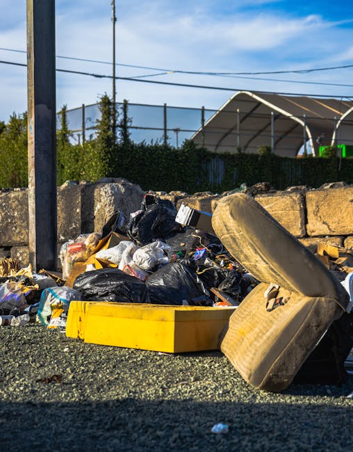 A close-up view of a rubbish pile situated outdoors on a gravel surface, featuring black plastic trash bags, discarded cardboard boxes, and various loose waste materials. In the foreground, a partially crushed and stained beige car tire leans against the debris, while a yellow plastic bin lies on its side amidst the refuse. Behind the pile, a low stone wall constructed from irregularly shaped concrete blocks separates the area from a background environment that includes a fence with green foliage, a tennis court with a metal fence and netting, and power lines overhead against a partly cloudy sky. The scene appears to depict an informal waste collection or on-site clearance activity, possibly linked to private rubbish removal services such as those offered by Rubbish Removal Croydon, as part of alternative rubbish handling solutions outside of municipal collections. The overall atmosphere is bright and clear, emphasizing the outdoor setting and the contrast between the waste materials and the surrounding environment.